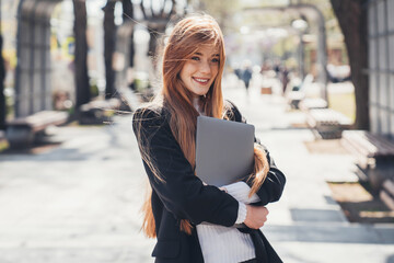 Caucasian ginger businesswoman in stylish clothes with a laptop walking outdoors in the park. Remote business. Attractive beautiful girl. Remote work.