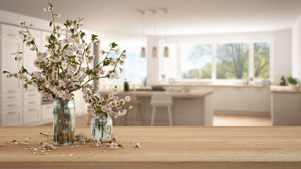 Wooden table, desk or shelf close up with branches of cherry blossoms in glass vase over blurred view of scandinavian white kitchen with island, boho interior design concept