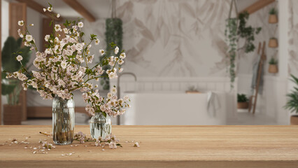 Wooden table, desk or shelf close up with branches of cherry blossoms in glass vase over blurred view of bohemian bathroom with bathtub, boho interior design concept