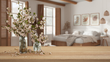 Wooden table, desk or shelf close up with branches of cherry blossoms in glass vase over blurred view of bohemian bedroom with double bed, boho interior design concept