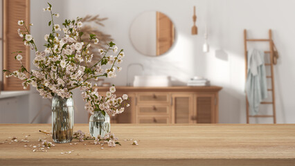 Wooden table, desk or shelf close up with branches of cherry blossoms in glass vase over blurred view of bohemian bathroom with washbasin, boho interior design concept