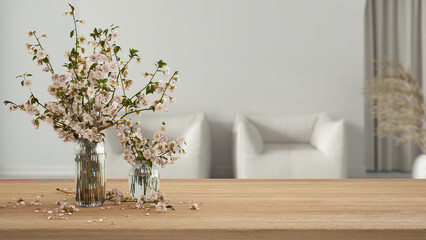 Wooden table, desk or shelf close up with branches of cherry blossoms in glass vase over blurred view of classic living room with armchairs, boho interior design concept