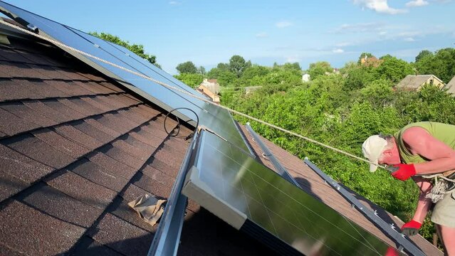 Worker installs solar panel on roof of small home secured with rope. DIY PV panels installation on bituminous tiles roof