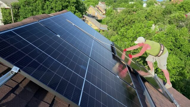 Installation process of PV solar panels on bituminous tiles roof of small house. Clean green renewable energy DIY concept. Worker secured with rope
