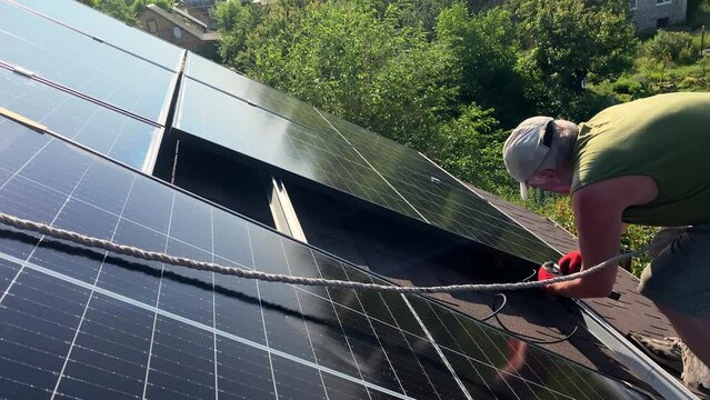 Worker fasten bolt installs solar PV panels on bituminous tiles roof of small house. DIY concept green energy