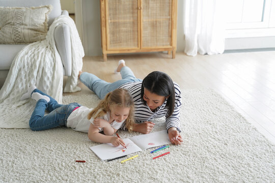 Carefree woman and little girl lying on floor together and painting with colorful pencils.