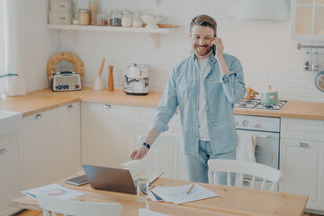 Handsome smiling bearded man speaking on cellphone in kitchen while working remotely
