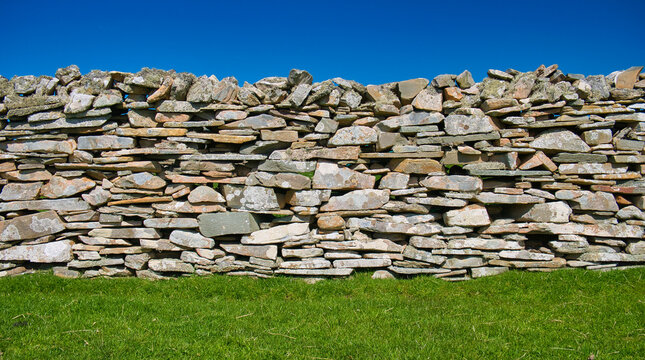 A colourful dry stone wall on a sunny day between a clear blue sky and green grass.
