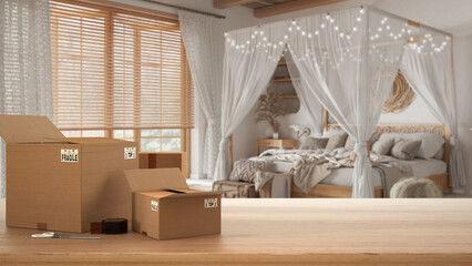 Wooden table, desk or shelf with stack of cardboard boxes over blurred view of bsohemian bedroom in boho style, interior design, moving house concept with copy space