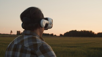 Fototapeta premium Farmer in virtual reality helmet in front of a sunset agricultural landscape. Man in a countryside field. Country life, food production, farming and technology concept