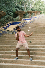 Obraz premium Young latin guy juggling in the sunny streets of Barcelona. Doing a show with some white balls while smiling. Dressed in brown pants and a pastel striped t-shirt.