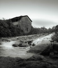 Grayscale shot of flooding water on rocky mud by an abandoned house with trees
