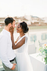 Smiling bride touches groom face with hands on terrace at home