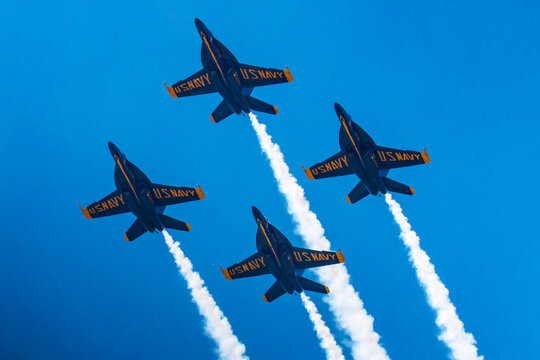 Four US Navy Blue Angel F18s flying in a diamond formation with a vapor trail