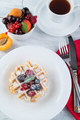 Blurred image of Belgian waffles with powdered sugar and fruit in a plate, cutlery, a cup of coffee and a plate of fruit.