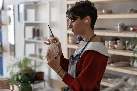 Happy concentrated woman painting clay jug with colourful paint shaping and sculpturing earthenware cup in pottery studio on ceramic workshop. Girl artist in earphones relaxing with art leisure - Powered by Adobe