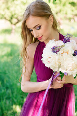 Fototapeta premium Portrait of a beautiful girl of model appearance in a purple fashion dress and a bouquet of peonies and lupines. Professional bridesmaid makeup. High quality photo.