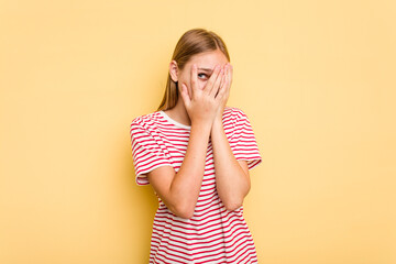 Young caucasian girl isolated on yellow background blink at the camera through fingers, embarrassed covering face.