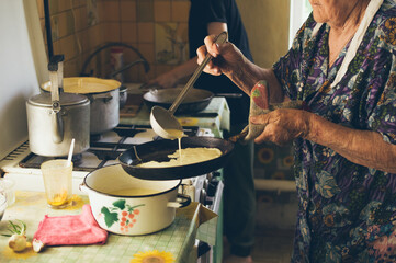 Old grandmother pours fluid dough to pan for  baking pancakes