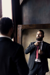 An attractive young man with a beard adjusts his tie in the mirror