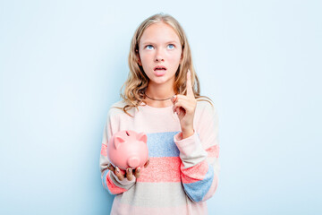 Caucasian teen girl holding a piggybank isolated on blue background pointing upside with opened mouth.