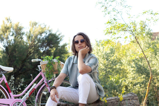 Young hansome long haired man on sunglasses sitting on a trunk looking to camera with a pink bike behind