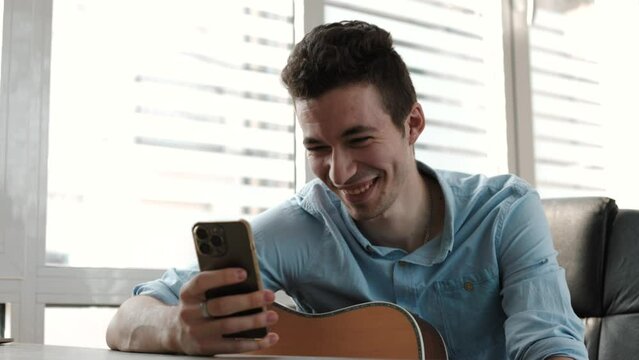 Young guitar player taking a break resting from playing string instrument using cellphone smiles read pleasant message. Hobby, pastime, modern technology