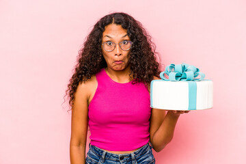 Young caucasian woman holding a cake isolated on pink background shrugs shoulders and open eyes confused.