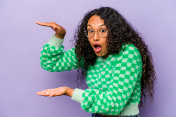 Young hispanic woman isolated on purple background shocked and amazed holding a copy space between hands.