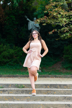 Chica joven guapa con gafas negras que esta posando delante de un monumento con vestido rosa manos en las caderas sonriendo y con pelo suelto casta&ntilde;o