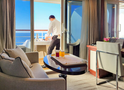 A room service waiter wearing a surgical mask brings a meal to a cruise ship cabin and balcony with the blue sea in view.