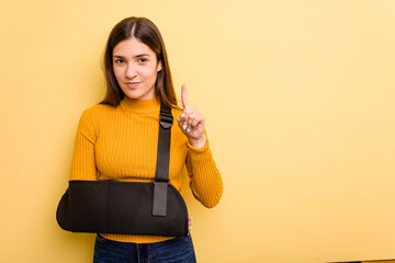 Young caucasian woman with broke arm isolated on yellow background showing number one with finger.
