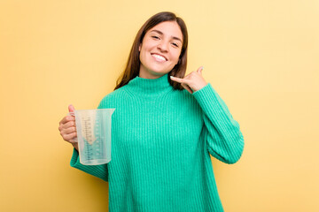 Young caucasian woman holding measuring jug isolated on yellow background showing a mobile phone call gesture with fingers.