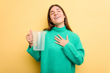 Young caucasian woman holding measuring jug isolated on yellow background laughs out loudly keeping hand on chest.