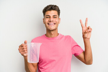 Young caucasian man holding measuring jug isolated on white background joyful and carefree showing a peace symbol with fingers.