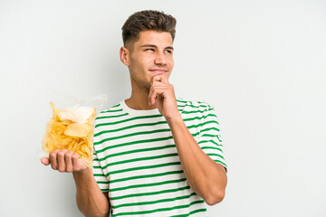 Young caucasian man holding crisps isolated on white background looking sideways with doubtful and skeptical expression.