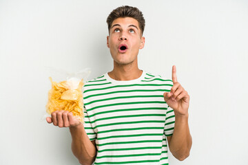 Young caucasian man holding crisps isolated on white background pointing upside with opened mouth.