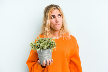 Young caucasian woman holding a plant isolated on blue background confused, feels doubtful and unsure.