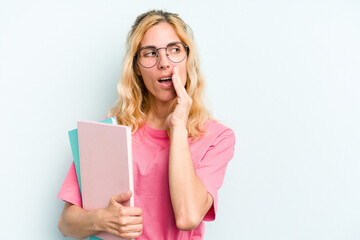 Naklejka premium Young student caucasian woman holding books isolated on blue background is saying a secret hot braking news and looking aside