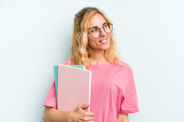 Young student caucasian woman holding books isolated on blue background looks aside smiling, cheerful and pleasant.