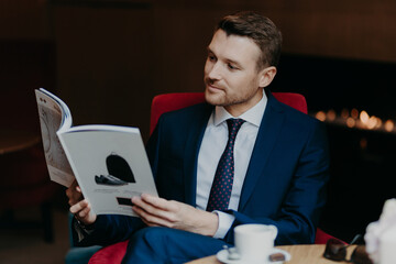 Fototapeta premium Horizontal shot of attractive male manager dressed in black suit, white shirt and tie, reads magazine in coffee shop, drinks hot beverage, has serious concetrated look. Business and leisure concept
