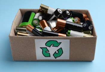 Used batteries in cardboard box with recycling symbol on light blue background, closeup