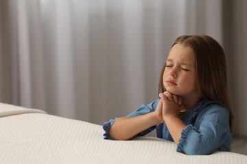 Naklejka premium Cute little girl with hands clasped together saying bedtime prayer in bedroom. Space for text