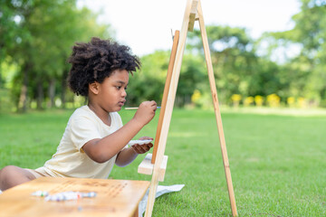 African American little boy with curly hair painting watercolor on white paper in summer park.Kid...