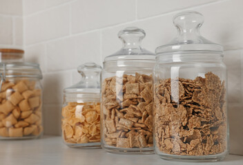 Glass containers with different breakfast cereals on white countertop near brick wall
