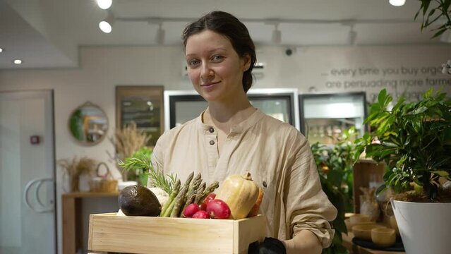 Woman with blue eyes looks at camera and smiles holding wooden box with fresh vegetables. Young woman with plait works in organic products shop