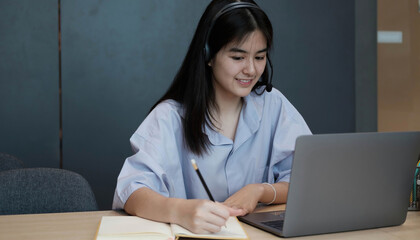 Smiling Asian young female using headset looking at laptop screen listen and learning online courses. Happy chinese business woman with headphones video call for customer service