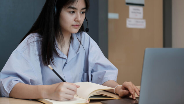 Smiling Asian young female using headset looking at laptop screen listen and learning online courses. Happy chinese business woman with headphones video call for customer service