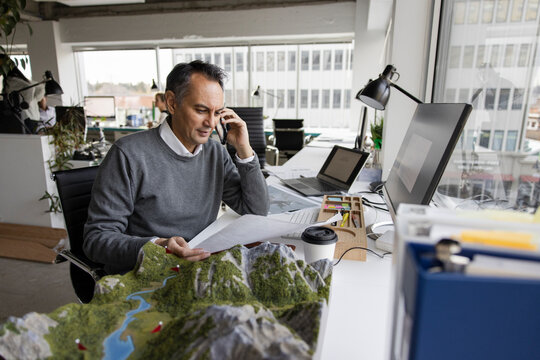 Male Architect Talking On Smart Phone At Desk With Landscape Model