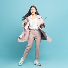 Young beautiful asian woman with smart casual cloth wearing pink coat smiling holding smartphone and laptop isolated on blue background